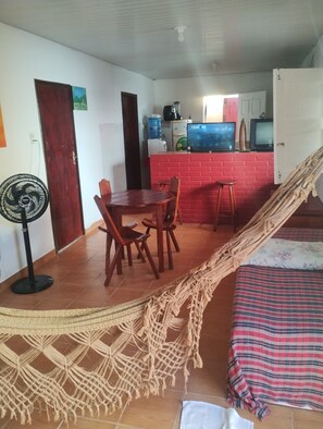Ein Zimmer mit Esstisch, Stühlen, einer Hängematte, einem Bett mit gestreiftem Überwurf, einem Ventilator und einer Küchenzeile mit roter Backsteinwand.