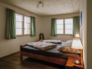 Et værelse med en træseng, to vinduer med grønne gardiner, en lampe på et natbord og et loft med en lysarmatur.