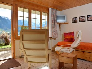 A room with a wooden chair, a bed with an orange blanket, a television on a stand, and a window with a mountain view. - Marielle Appartement/Fewo, Dusche, WC, 1 Schlafraum 2-Zimmerwohnung by Interhome (Adelboden)