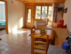 Ein gemütliches Zimmer mit einem Holztisch, Stühlen, einer Couch, einem Fernseher und einem Fenster mit Vorhängen.