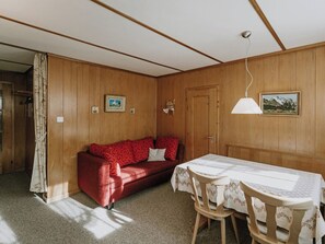 Ein gemütliches Zimmer mit einer Holzwand, einem roten Sofa, einem Esstisch mit Stühlen und einer hängenden Lampe.