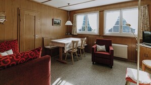 Una habitación acogedora con paredes de madera, una mesa de comedor con sillas, un sillón rojo, una mesita con una lámpara y una ventana con cortinas.