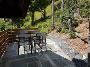 Eine sonnige Terrasse mit Steintisch und -stühlen, umgeben von einer Steinmauer und viel Grün.