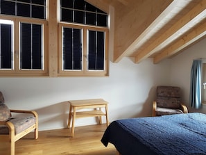 Ein Zimmer mit einem Bett, zwei Stühlen, einem kleinen Tisch und großen Fenstern mit Jalousien.