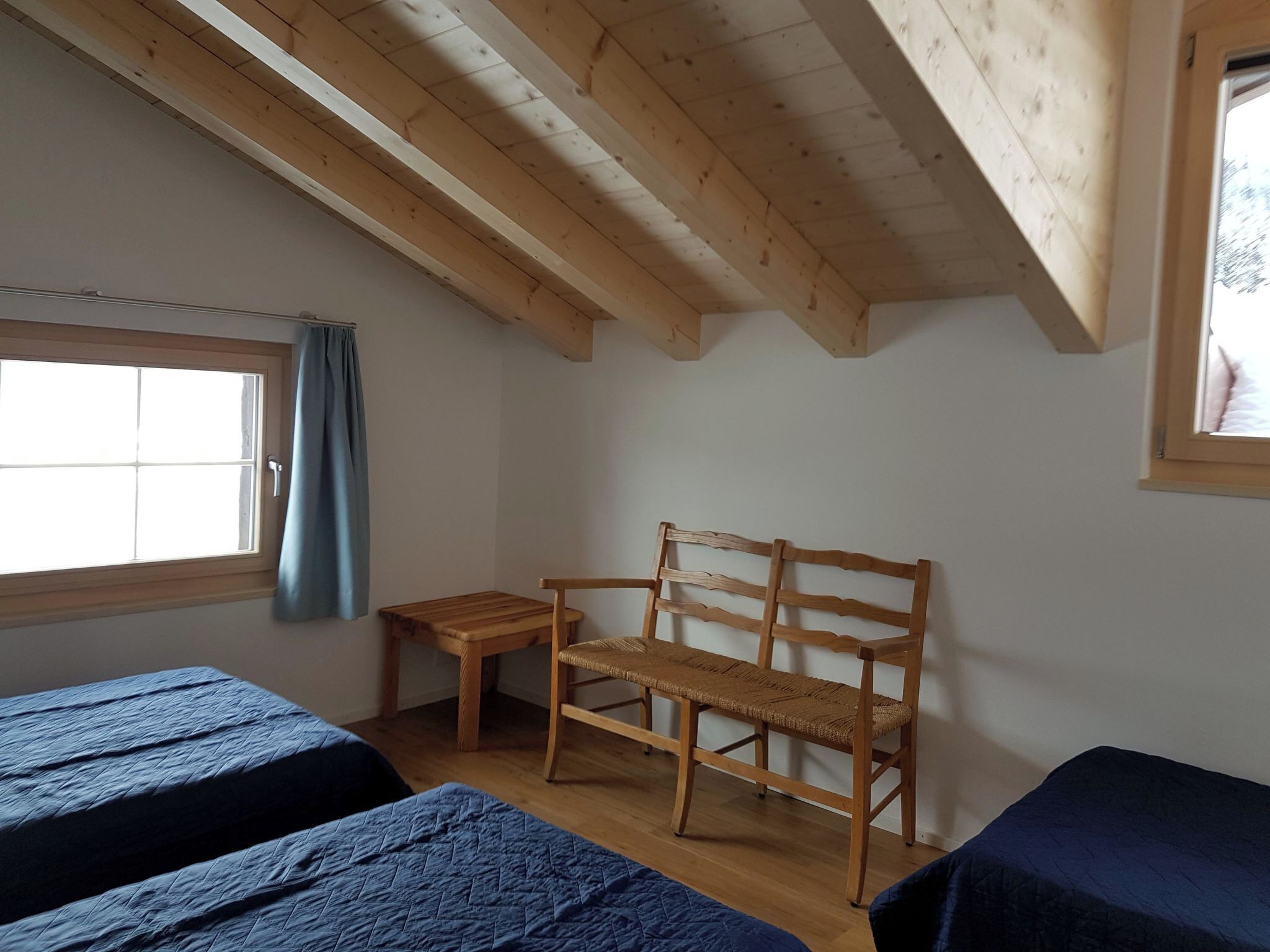 A room with two beds, a wooden bench, a small table, and a window with a curtain.