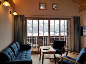 Une pièce avec une grande fenêtre, un canapé, un fauteuil, une table et un téléviseur fixé au mur.