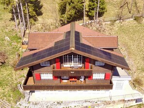 A large house with a sloped roof, solar panels, a covered patio with outdoor seating, and a view of a hilly landscape. - Chalet Ginette Appartement/Fewo, Bad, WC, 2 Schlafräume by Interhome (Adelboden)