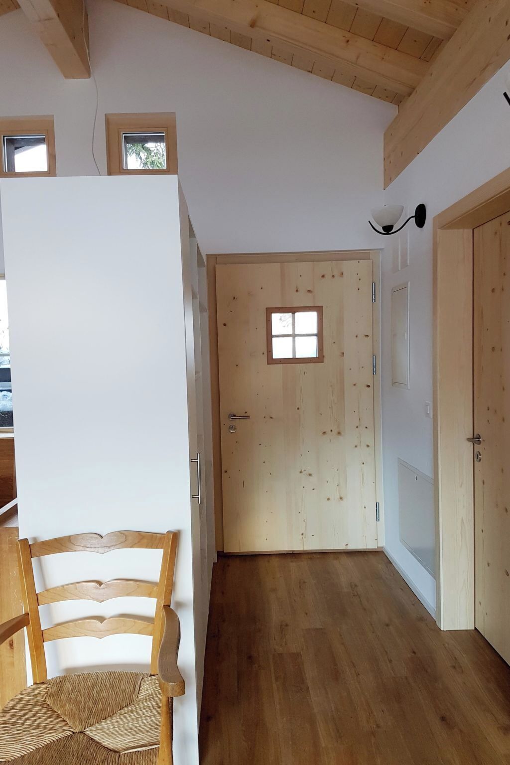 Un couloir doté d’un plancher en bois, une porte en bois avec une fenêtre, et une chaise avec un siège en osier.