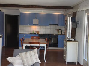 Een functionele keuken met blauwe kastjes, een eettafel met stoelen, een bank met kussens, en een klein wit kastje.