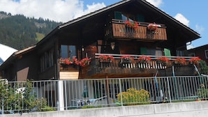 Ein zweistöckiges Holzhaus mit Balkonen, einem Metallzaun im Vordergrund und einer Bergkulisse.