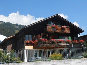 Ein zweistöckiges Holzhaus mit Balkonen, einem Metallzaun im Vordergrund und einer Bergkulisse.