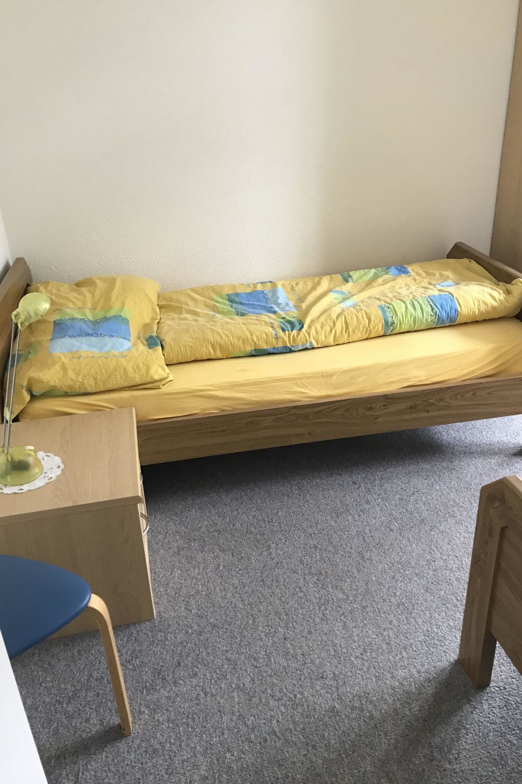 Een eenpersoonsbed met een deken met een geel en blauw patroon, een bedlampje en een houten bureau met een stoel.
