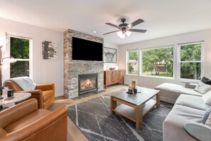 A modern living room with a fireplace, comfortable seating, a flat-screen TV, and large windows offering a view of the outdoors. - Whitefish Mountain Retreat with Hot Tub, Fire Pit & Heated Garage (Whitefish)