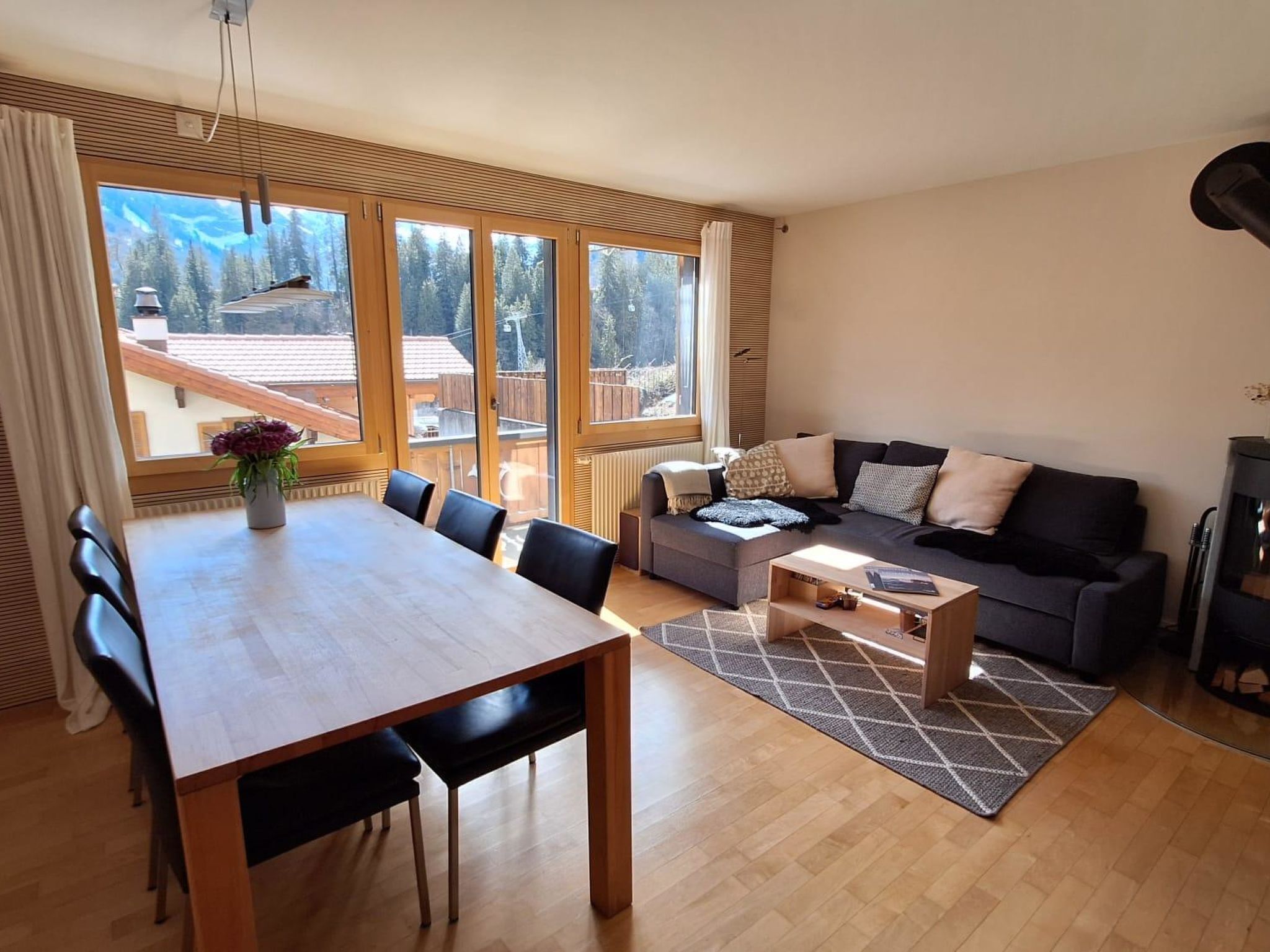 Un salon moderne avec une table à manger en bois, des chaises noires, un canapé gris et une table basse. De grandes fenêtres offrent une vue sur une terrasse en bois et des arbres.