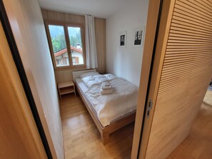 A small, single-bed room with a wooden floor, a window with curtains, and a view of a building with a red roof. - Chalet Sirius Appartement/Fewo, Bad, WC, 3 Schlafräume 4-Zimmer-Wohnung by Interhome (Adelboden)