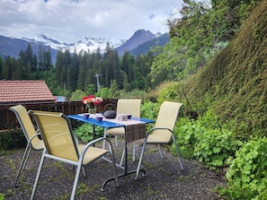 An outdoor seating area with a table and chairs, surrounded by greenery and a mountainous backdrop. - Chalet Sirius Appartement/Fewo, Bad, WC, 3 Schlafräume 4-Zimmer-Wohnung by Interhome (Adelboden)