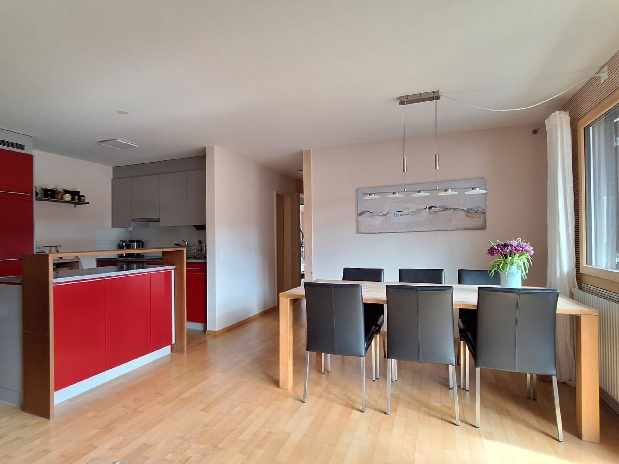 Een moderne keuken met rode kastjes, een eethoek met een houten tafel en zwarte stoelen, en een groot raam met uitzicht.