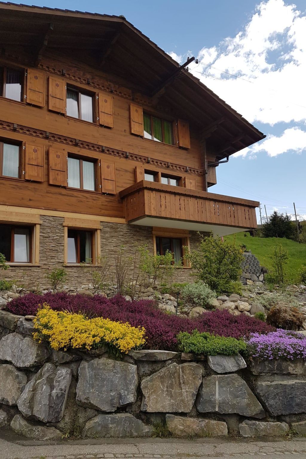 Un edificio de madera de varios pisos con fachada de piedra, balcones y un jardín ajardinado con diversas plantas.