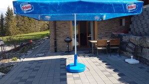 A patio area with a blue umbrella branded with Nestle Frisco, a stone wall, and a wooden table with chairs. - Grüneggli Appartement/Fewo, Bad, WC, 2 Schlafräume 3.Zimmerwohnung by Interhome (Adelboden)