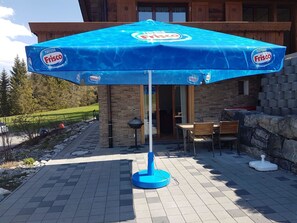 A patio area with a blue umbrella branded with Nestle Frisco, a stone wall, and a wooden table with chairs. - Grüneggli Appartement/Fewo, Bad, WC, 2 Schlafräume 3.Zimmerwohnung by Interhome (Adelboden)