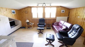 Ein Zimmer mit Holzboden, einem Ledersofa, einem Ledersessel, einem kleinen Tisch mit Lampe sowie einem Fernseher auf einem Beistelltisch.