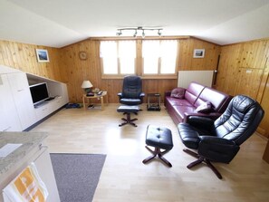 Ein Zimmer mit Holzboden, einem Ledersofa, einem Ledersessel, einem kleinen Tisch mit Lampe sowie einem Fernseher auf einem Beistelltisch.