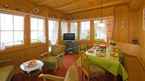 Een kamer met houten lambrisering, een eettafel gedekt voor vier personen, stoelen, een televisie en grote ramen.