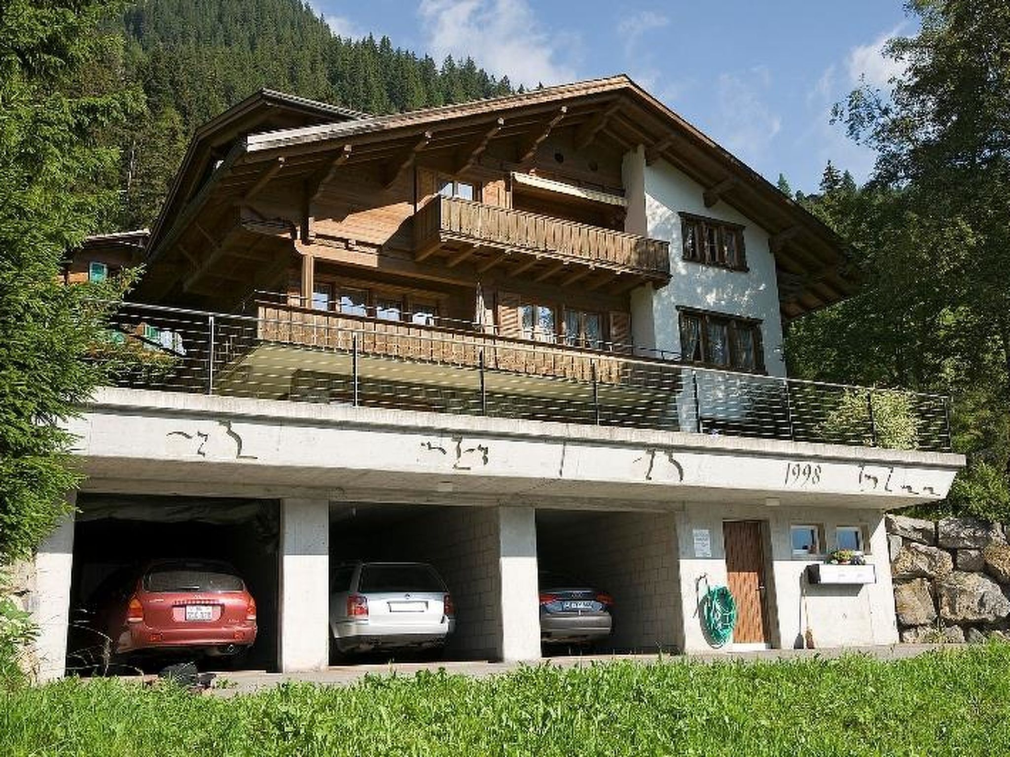 Ein zweigeschossiges Gebäude mit Balkon, ein Parkplatz mit drei Autos und eine gebirgige Kulisse.
