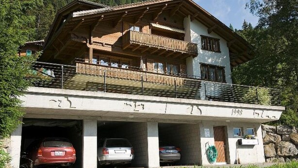 Een gebouw met twee verdiepingen, een balkon, parkeergelegenheid onder een overkapping en een bergachtige achtergrond.