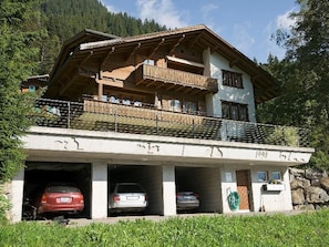 Ein zweistöckiges Gebäude mit Balkon, überdachtem Parkplatz und Bergelandschaft im Hintergrund.