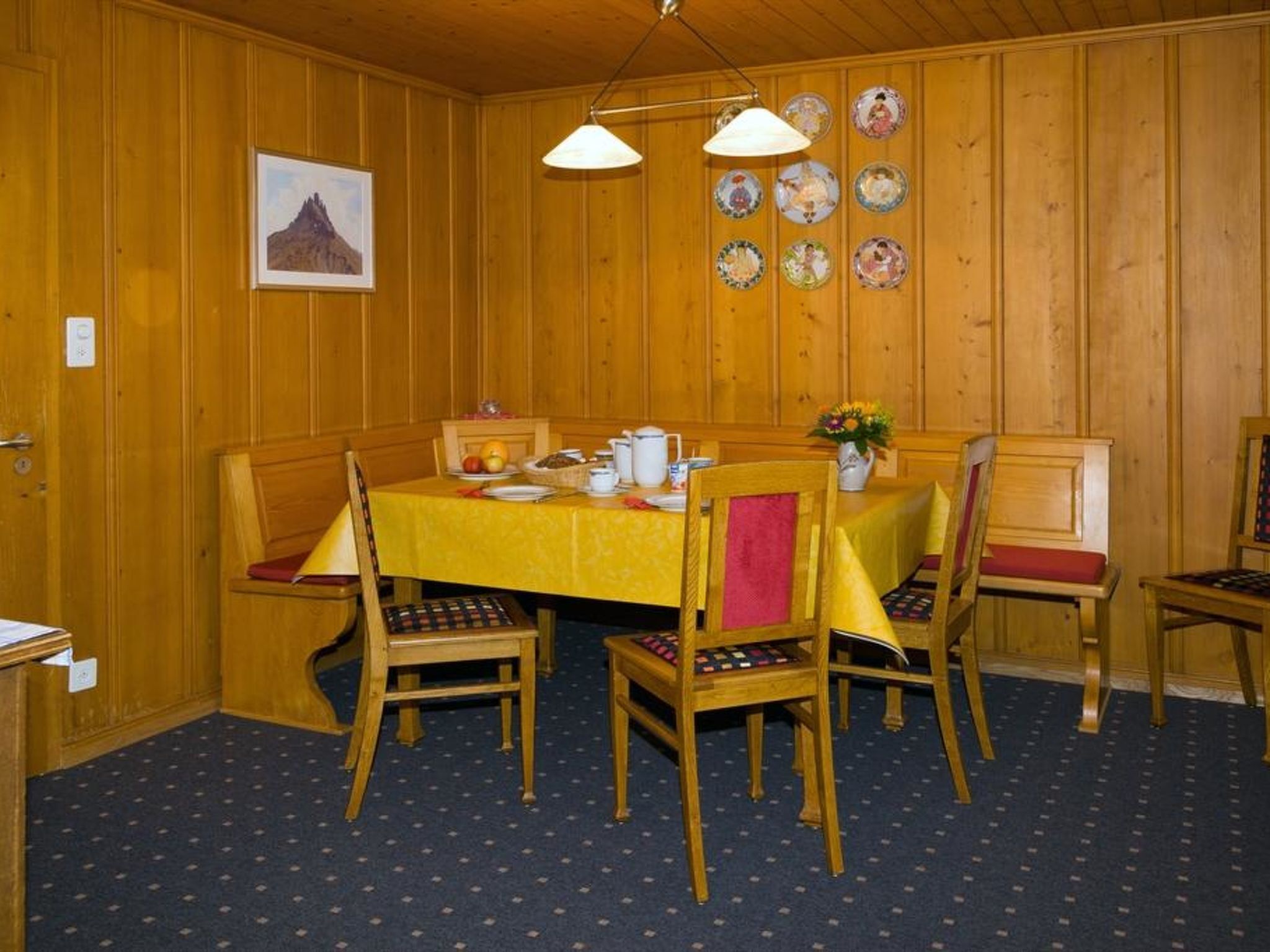 Puisilla paneeleilla verhoiltu ruokasali, jossa on neljälle katettu ruokapöytä, pehmustetuilla istuimilla varustetut tuolit, seinällä taulu ja seinällä esillä oleva lautaskokoelma.