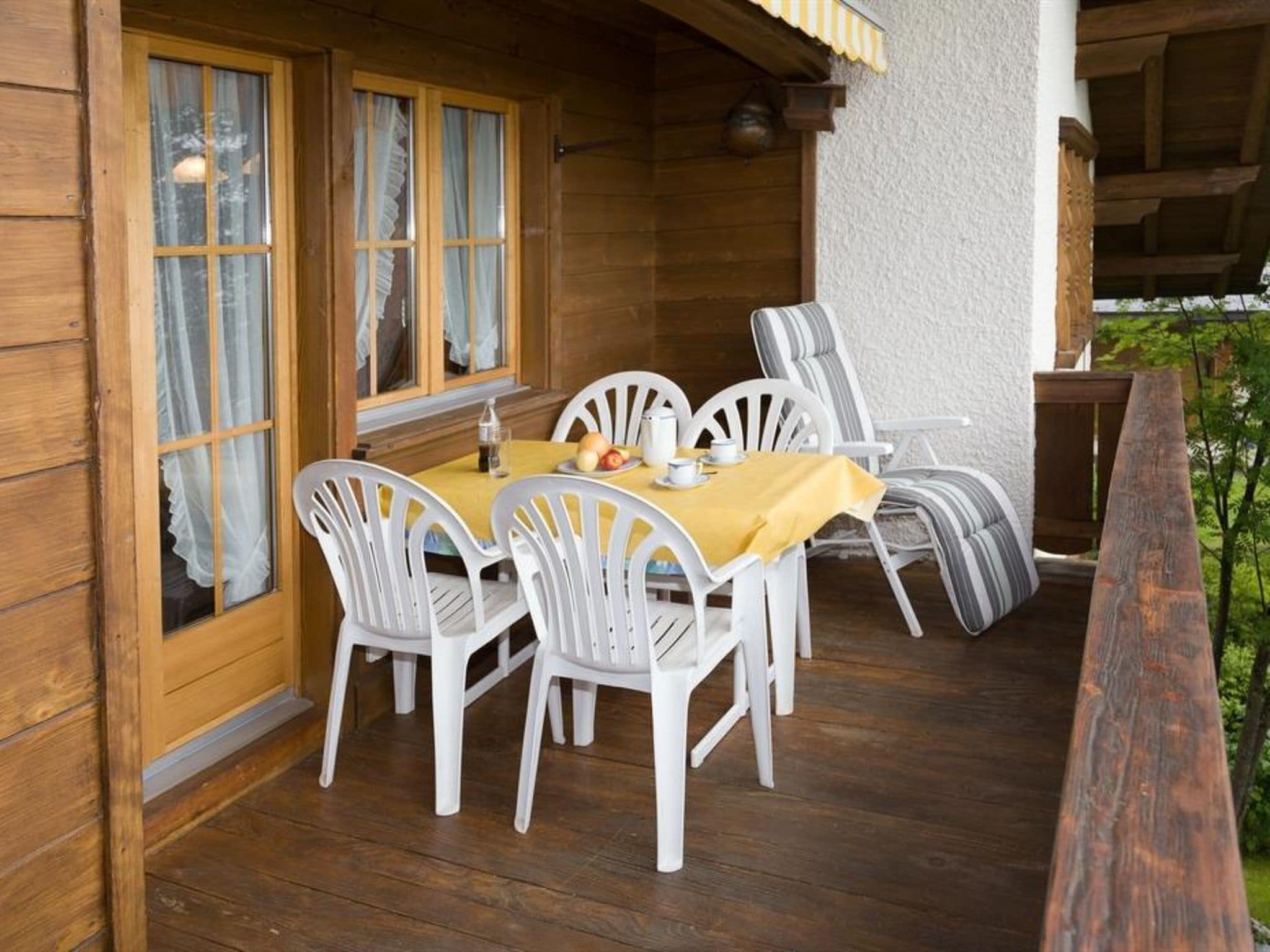 A wooden deck with a table and chairs, a window with curtains, and a view of greenery.