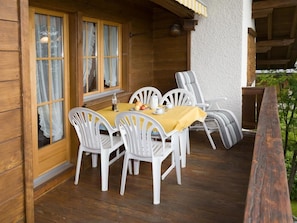 Eine Holzterrasse mit Tisch und Stühlen, ein Fenster mit Vorhängen und Blick ins Grüne.