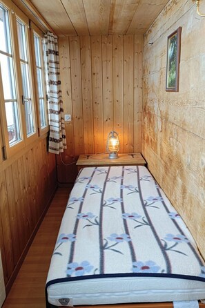 Ein Zimmer in einer Holzhütte mit einem Bett, einem Fenster mit Vorhängen, einer Wandleuchte und einem kleinen Bild.