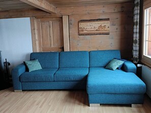 Ein blaues Ecksofa mit passenden Kissen, eine Holzwand mit einem aufgehängten Bild und ein Fenster mit gemusterten Vorhängen.