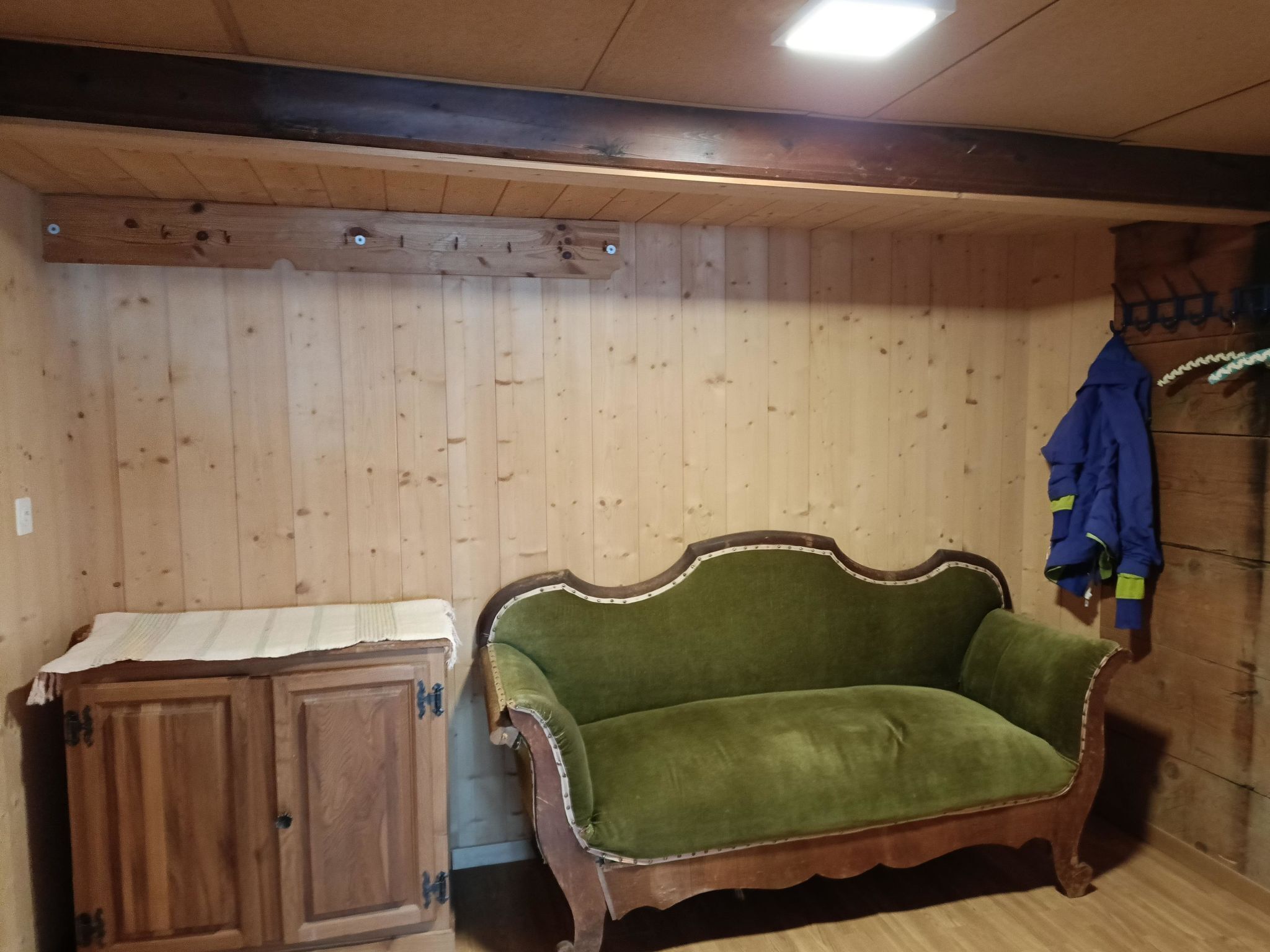A room with wooden panel walls, a green upholstered sofa, a wooden cabinet, and a coat hanging on the wall.