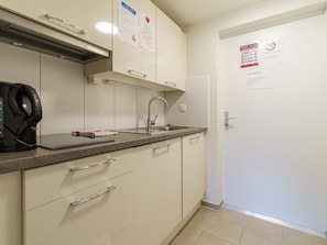 A kitchenette with a stove, sink, and cabinets. There are signs on the wall and a door with a notice. - Haus Astoria by Interhome (Saas-Grund)