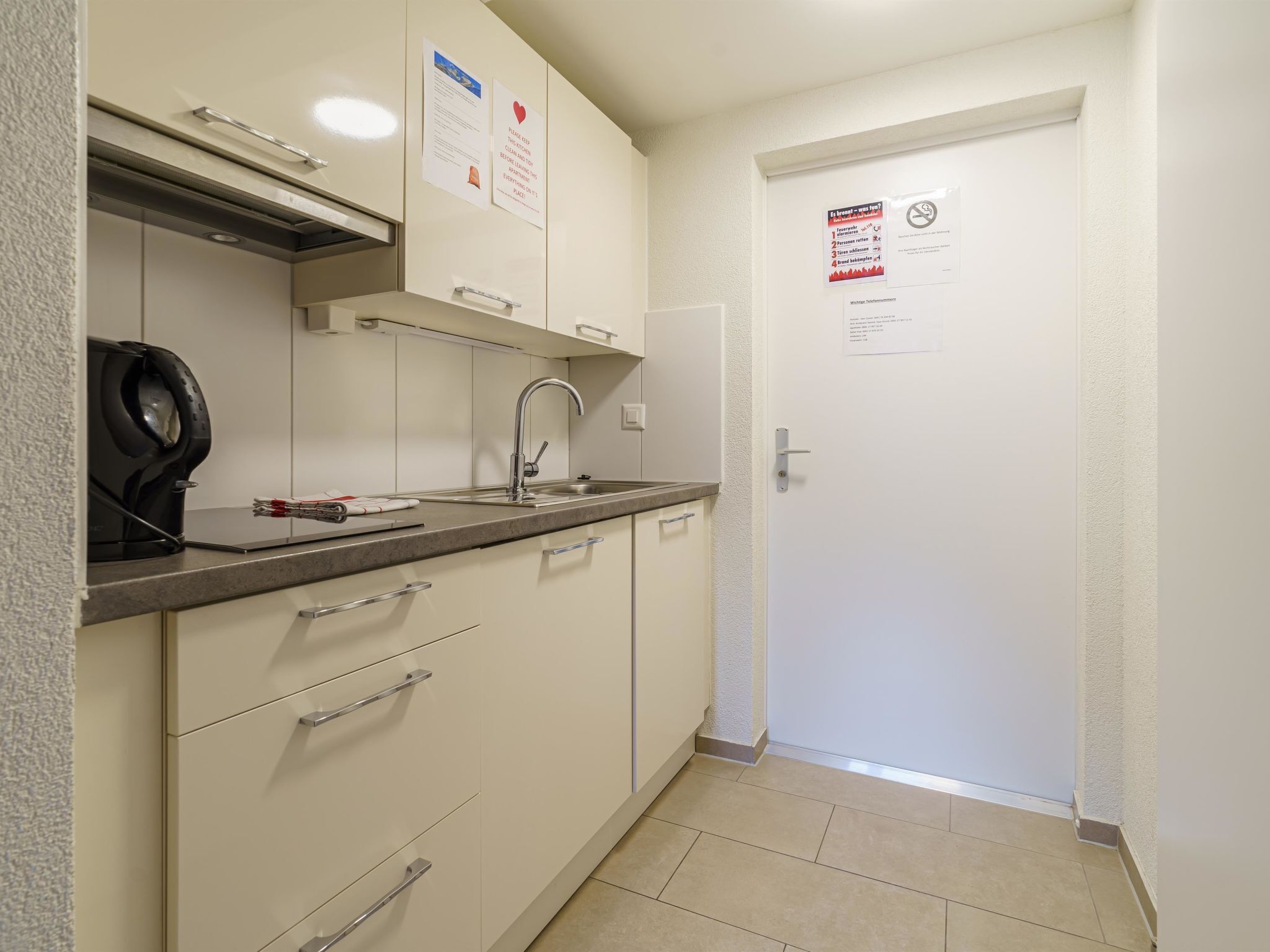 Een kleine keuken met een spoelbak, kookplaat en kastjes. Er hangen borden aan de muur en een deur met een bericht.