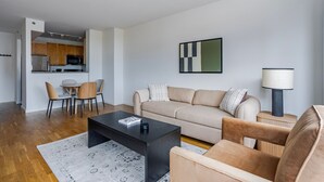Ein modernes Wohnzimmer mit Sofa, Couchtisch und Sesseln, ein Küchenbereich mit Schränken und Geräten sowie ein Esstisch mit Stühlen.