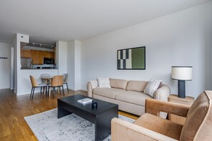 Ein modernes Wohnzimmer mit Sofa, Couchtisch und Sesseln, ein Küchenbereich mit Schränken und Geräten sowie ein Esstisch mit Stühlen.