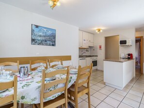 A dining area with a table set for four, wooden chairs, a painting of a mountain landscape, and a kitchen with white cabinets and a microwave. - Haus Alfa by Interhome (Saas-Fee)