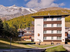 Ein modernes Gebäude mit der Aufschrift 'Alfa' auf der Fassade, umgeben von einer gebirgigen Landschaft mit schneebedeckten Gipfeln und einem bewaldeten Gebiet.