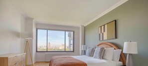 Ein Hotelzimmer mit großem Fenster, einem Bett mit Kopfteil, Nachttischlampen und Blick auf die Stadt.