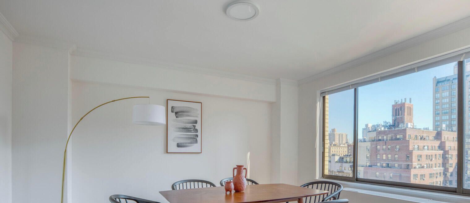 Ein Zimmer mit Esstisch, Stühlen, einer Vase, einer Stehlampe und einem gerahmten abstrakten Kunstwerk. Ein großes Fenster bietet Blick auf Gebäude.