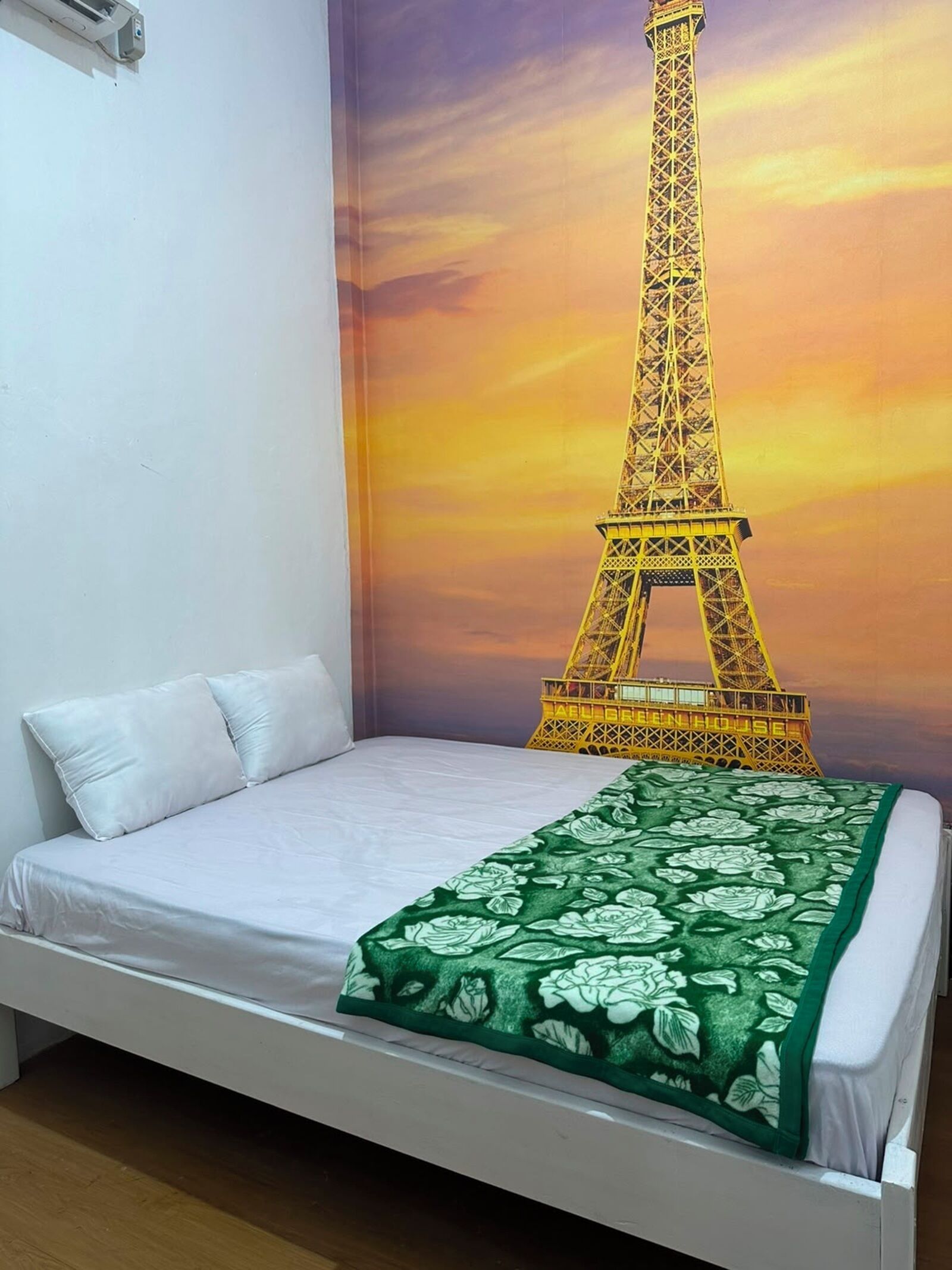 A bed with white bedding and a green floral throw, a white pillow, and a wall with a sunset mural of the Eiffel Tower.