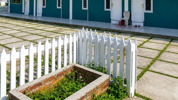 Un edificio de dos pisos con fachada verde y detalles blancos, que presenta una hilera de ventanas y puertas, una cerca blanca de piquetes y una jardinera de ladrillo con follaje.