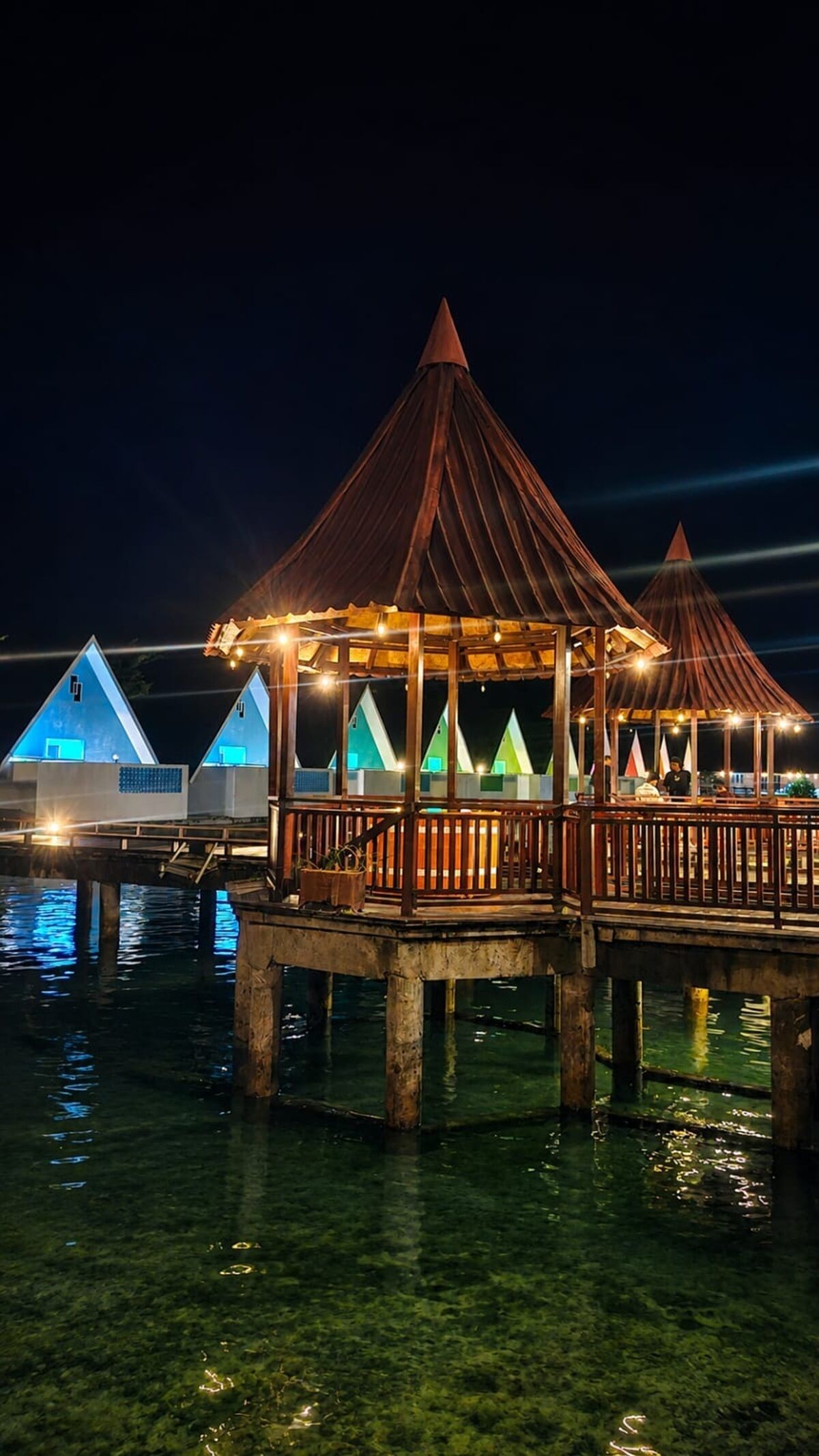 A wooden structure with a roof, surrounded by water, and adjacent to a building with a blue exterior.