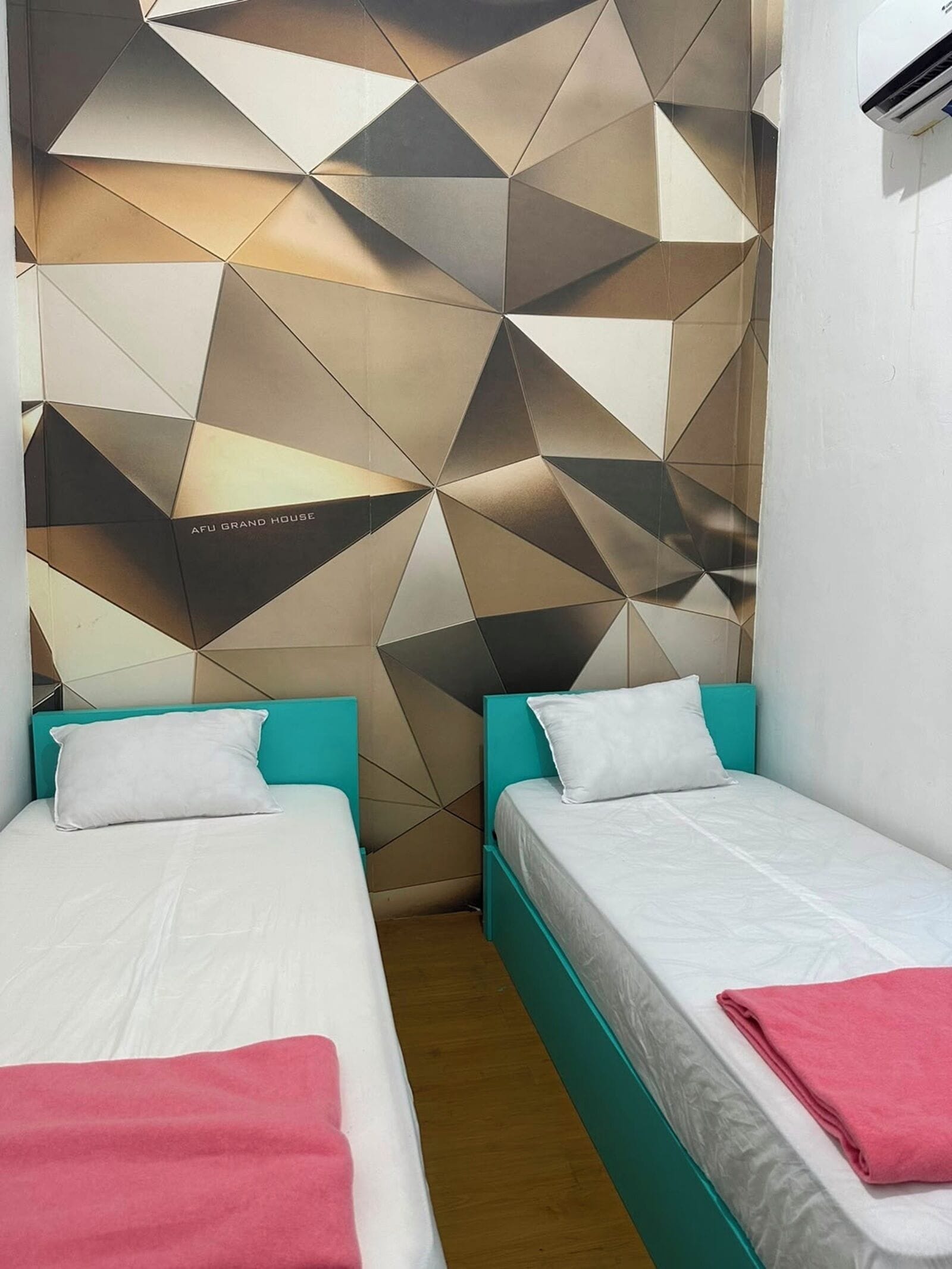 A hotel room with two beds, each with a white mattress and teal headboard. There is a geometric patterned wall behind the beds and a white air conditioning unit on the wall.