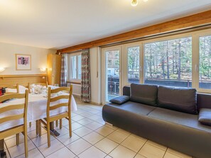 A room with a dining table set for four, a sofa, and large windows offering a view of a forested area. - Haus Alfa by Interhome (Saas-Fee)
