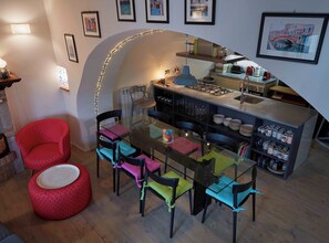 Een compact woongedeelte met een eethoek bestaande uit een glazen tafel en stoelen, een keuken met moderne apparatuur, en een gezellige zithoek met rode poefs.